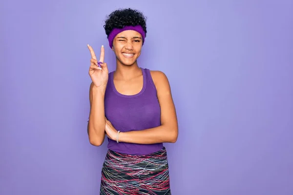 Beautiful african american afro woman wearing casual sportswear over purple background smiling with happy face winking at the camera doing victory sign. Number two.
