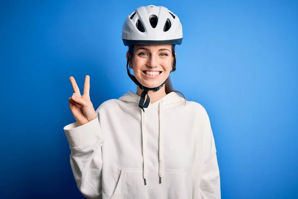 年轻美丽的红头发骑自行车的女人戴着自行车头盔 带着孤独的蓝色背景微笑着 脸上挂着喜庆的表情看着摄像机做胜利的标志 第二点 — 图库照片