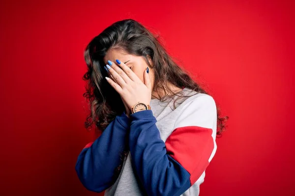 Young beautiful woman with curly hair wearing casual sweatshirt over isolated red background with sad expression covering face with hands while crying. Depression concept.