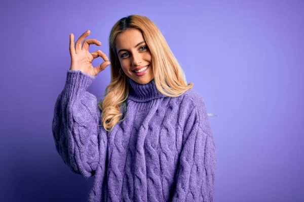 紫色の背景にカジュアルなタートルネックセーターを身に着けている若い美しいブロンドの女性は 手と指でOkサインをして肯定的な笑顔 成功した表現 — ストック写真