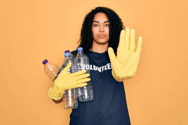 Young african american woman doing volunteering recycling holding plastic bottles with open hand doing stop sign with serious and confident expression, defense gesture