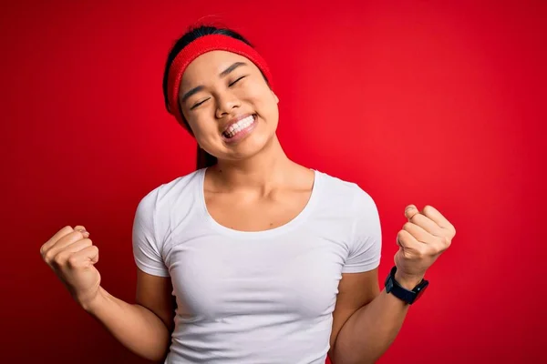年轻美丽的亚洲女运动员身穿运动服 在红色背景下做运动 非常高兴 兴奋地举起双臂做获胜者的手势 微笑着 尖叫着要成功 庆祝概念 — 图库照片