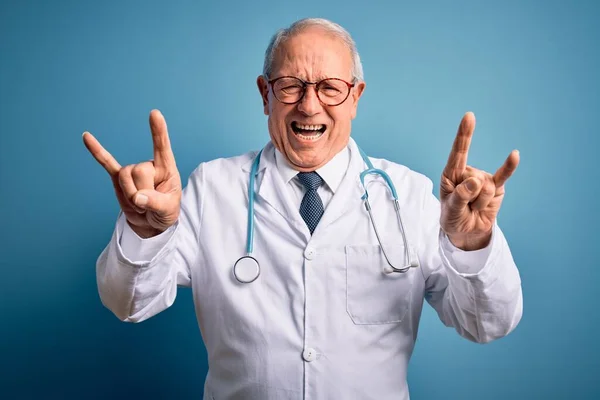 Senior grey haired doctor man wearing stethoscope and medical coat over blue background shouting with crazy expression doing rock symbol with hands up. Music star. Heavy concept.