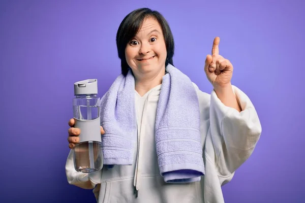 Young down syndrome fitness woman training at gym holding water bottle and sport towel surprised with an idea or question pointing finger with happy face, number one