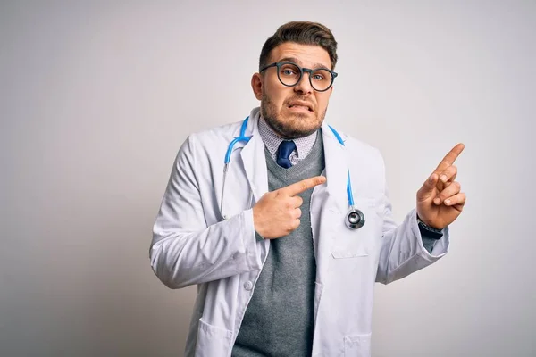 Young doctor man with blue eyes wearing medical coat and stethoscope over isolated background Pointing aside worried and nervous with both hands, concerned and surprised expression