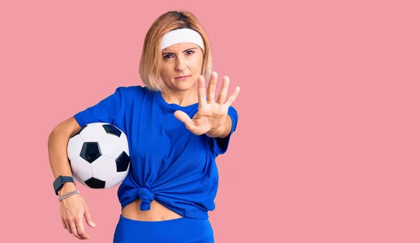 Young blonde woman holding soccer ball with open hand doing stop sign with serious and confident expression, defense gesture