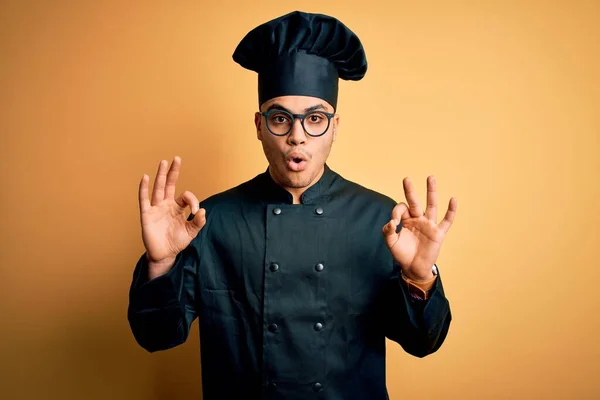 年轻的巴西厨师 身穿炊具制服 头戴黄底帽 看上去惊讶而震惊 用手指做着认可的标志 疯狂的表情 — 图库照片