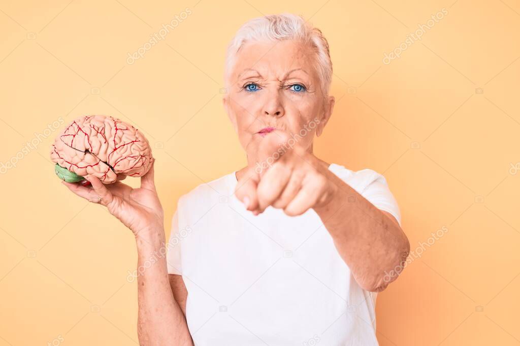 Senior beautiful woman with blue eyes and grey hair holding brain as mental health concept pointing with finger to the camera and to you, confident gesture looking serious 