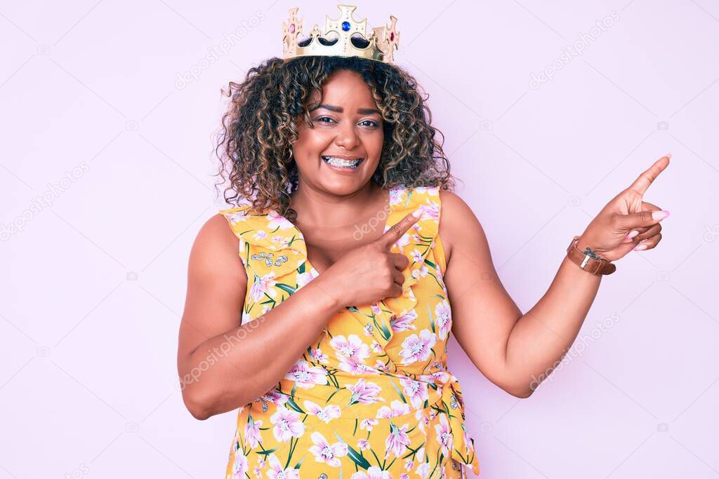 Young african american plus size woman wearing princess crown smiling and looking at the camera pointing with two hands and fingers to the side. 