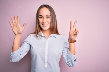 Zarif bir tişört giyen genç ve güzel bir iş kadını pembe arka planda duruyor ve yedinci parmaklarıyla işaret ederken kendinden emin ve mutlu bir şekilde gülümsüyor..