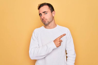 Mavi gözlü, sıradan bir süveter giyen, sarı arka planda duran, el parmağı ile reklamını yapan, ciddi ve sakin yüzlü genç bir adam.