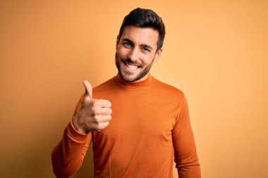 Sarı arka planda dikilmiş sakallı, genç ve yakışıklı bir adam elleriyle mutlu bir jest yapıyor. Kameraya bakıp başarıyı gösteren ifade onaylanıyor.