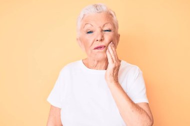 Son sınıf güzeli, mavi gözlü, gri saçlı, sarı arka planda klasik beyaz tişört giyen dişleri ağrıyan diş ağrısı ya da diş hastalığından dolayı ağızlarına acı dolu bir ifadeyle dokunan bir kadın. dişçi 