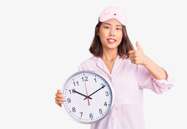 Genç güzel Çinli kız uyku maskesi takıyor ve pijama giyiyor. Elinde kocaman bir saat var. Mutlu ve pozitif gülümsüyor. 