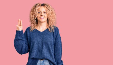 Kıvırcık saçlı, sıradan bir süveter giyen genç sarışın kadın üç numaralı parmaklarıyla onu gösterirken kendinden emin ve mutlu gülümsüyor.. 
