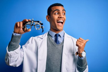 Yakışıklı Afro-Amerikan optik adam elinde optometri gözlüklerini mavi arka planda tutuyor ve baş parmağıyla gülümsüyor.