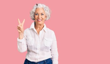 Kıdemli, gri saçlı, günlük kıyafetler giyen, kendini beğenmiş ve mutlu bir şekilde gülümseyen, iki numarayı işaret eden kadın.. 