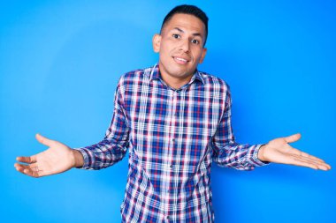 Gündelik kıyafetler giyen genç, yakışıklı Latin bir adam. Kolları ve elleri havada, kafası karışmış bir ifade. şüphe kavramı. 