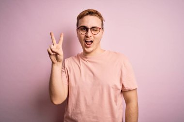 Genç, kızıl saçlı, günlük tişört giyen, pembe arka planda duran mutlu yüzüyle kameraya gülümseyen ve parmaklarıyla zafer işareti yapan bir adam. İki numara..