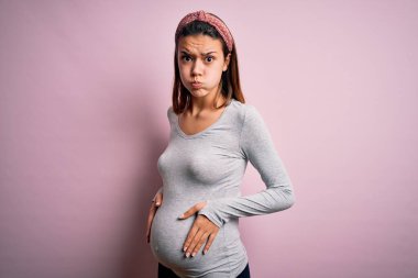 Young beautiful teenager girl pregnant expecting baby over isolated pink background puffing cheeks with funny face. Mouth inflated with air, crazy expression. clipart