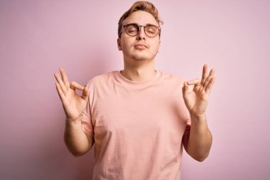 Genç, kızıl saçlı, günlük tişört giyen, izole edilmiş pembe arka plan üzerinde duran, gözleri kapalı, parmaklarıyla meditasyon hareketi yapan yakışıklı bir adam. Yoga konsepti.