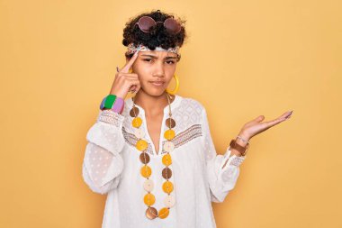 Güneş gözlüğü ve aksesuar takan Afro-Amerikan Afro-Amerikan genç bir kadın. Kafası karışmış ve avuç içi açık bir şekilde fotokopi alanı gösterip alnını işaret ediyor. Bunu bir düşün..