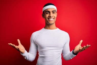 Genç ve yakışıklı Afro-Amerikan sporcusu kırmızı arka planda spor giysisi giyiyor. Neşeli bir şekilde kollarını açıp gülümsüyor. Dostça hoş geldin, olumlu ve kendinden emin selamlar.