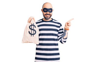Hırsız maskesi takan genç yakışıklı bir adam dolar sembollü para çantası tutuyor mutlu bir şekilde elini ve parmağını yana doğru uzatıyordu. 