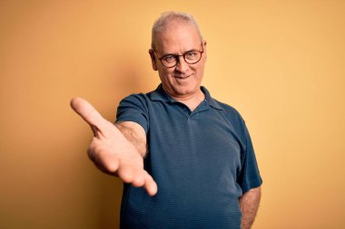 Orta yaşlı yakışıklı beyaz adam sarı arka planda günlük polo ve gözlük takıyor. Gülümsüyor, gülümsüyor, yardım ve kabullenme eli uzatıyor..