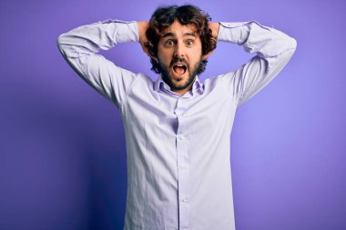 Mor arka plan üzerinde duran sakallı, genç ve yakışıklı iş adamı elleri kafasında çılgın, korkmuş ve ağzı açık şoktan şaşırmış.