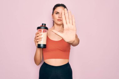 Spor kıyafetleri giyen genç ve güzel bir kadın. Açık elle protein içeceği içiyor. Dur işareti yapıyor.
