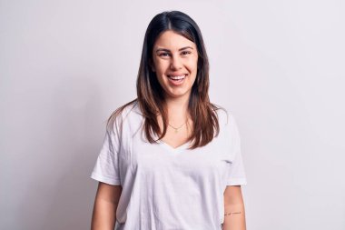 Genç, güzel, esmer, günlük tişörtler giyen bir kadın izole edilmiş beyaz arka planda mutlu ve havalı bir gülümsemeyle dikiliyor. Şanslı kişi..