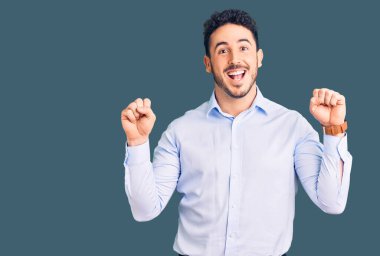 İş kıyafetleri giyen genç İspanyol adam şaşırmış ve şaşırmış bir şekilde kollarını kaldırmış ve gözlerini açmış bir şekilde başarıyı kutluyor. Kazanan konsept. 