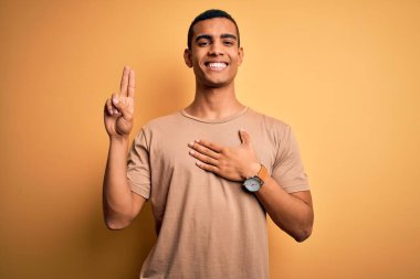 Young handsome african american man wearing casual t-shirt standing over yellow background smiling swearing with hand on chest and fingers up, making a loyalty promise oath clipart