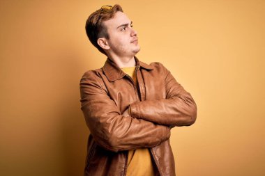 Genç, yakışıklı, deri ceket giyen, izole edilmiş sarı arka planda kolları çapraz ve kendinden emin genç bir adam.