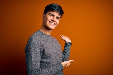 İzole edilmiş turuncu arka planda duran sıradan süveter giyen genç yakışıklı adam gülümseyerek içeri girmeye davet ediyor.