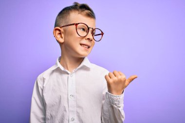 Mavi gözlü, mor arka planda beyaz tişörtlü, mutlu yüzüyle gülümseyen ve baş parmağıyla yüzünü gösteren beyaz bir çocuk..