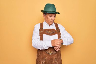 Sarı arka planda Alman geleneksel Ekim Festivali kıyafeti giyen yaşlı gri saçlı adam kol saatindeki saati kontrol ediyor, rahat ve kendinden emin.