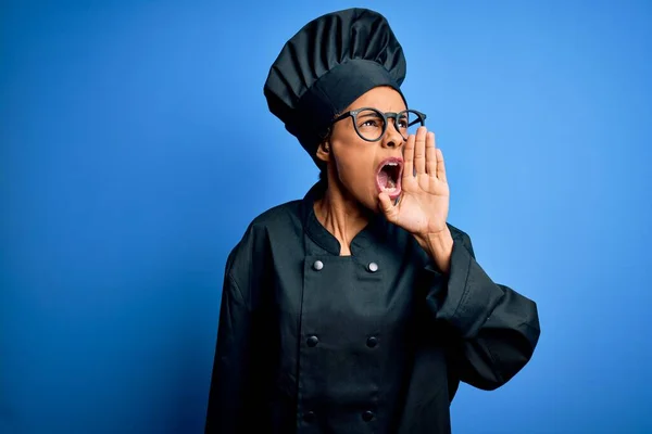 Young african american chef woman wearing cooker uniform and hat over blue background shouting and screaming loud to side with hand on mouth. Communication concept.