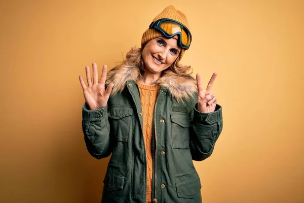 中年漂亮的金发滑雪者女人穿着雪地运动鞋和滑雪护目镜 带着七号手指指尖 面带微笑 自信而快乐 — 图库照片