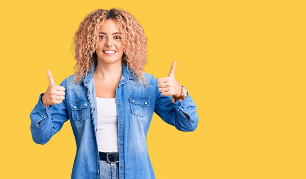 Young blonde woman with curly hair wearing casual denim jacket success sign doing positive gesture with hand, thumbs up smiling and happy. cheerful expression and winner gesture.