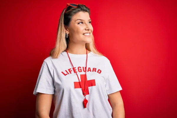 Young beautiful blonde lifeguard woman wearing t-shirt with red cross and whistle looking away to side with smile on face, natural expression. Laughing confident.