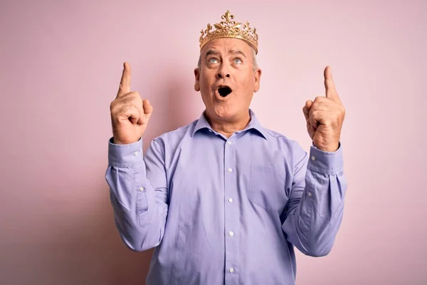 中年ホーリーマン身に着けています黄金の冠を身に着けていますキングスタンド上の隔離されたピンクの背景驚きと驚きを見上げ指と上げ腕 — ストック写真