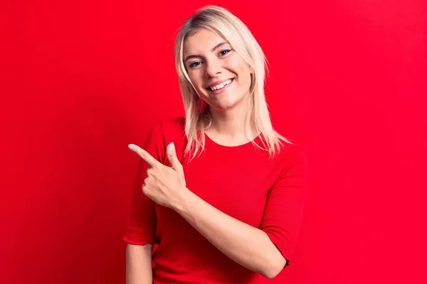 年轻美丽的金发女人穿着休闲的红色T恤 站在孤零零的背景上 高兴地微笑着 手指头指向旁边 — 图库照片