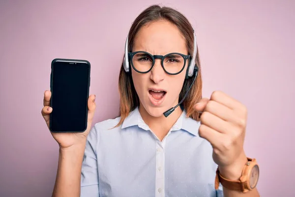 Beautiful call center agent woman working using headset holding smartphone holding screen annoyed and frustrated shouting with anger, crazy and yelling with raised hand, anger concept