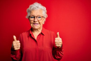 Son sınıf güzeli gri saçlı kadın kırmızı arka plan işareti üzerine günlük gömlek ve gözlük takıyor. Elleriyle olumlu jest yapıyor. Gülümsüyor ve mutlu. Neşeli ifade ve kazanan jest.