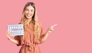 Güzel, beyaz, sarı saçlı, kalp takvimiyle gülümseyen, elini ve parmağını yana doğru gösteren bir kadın. 