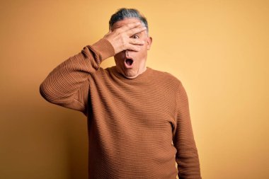 Orta yaşlı, yakışıklı, gri saçlı, sarı arka planda sıradan bir süveter giyen, yüzünü ve gözlerini kapayan, utangaç bir ifadeyle parmaklarından bakan bir adam..