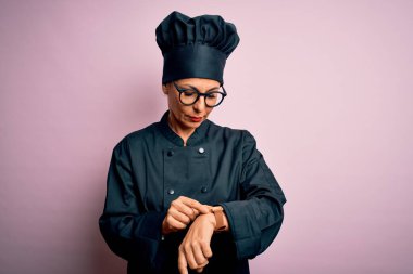 Orta yaşlı, esmer, aşçı üniforması giyen ve şapkalı bir kadın. İzole edilmiş pembe bir geçmişi var. Saati kontrol ediyor, rahat ve kendinden emin.