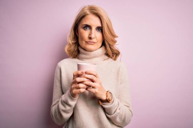 Orta yaşlı, sarışın, pembe kahve fincanı içen güzel bir kadın izole bir arka planda kendine güvenen bir yüz ifadesiyle ciddi ciddi düşünüyor.